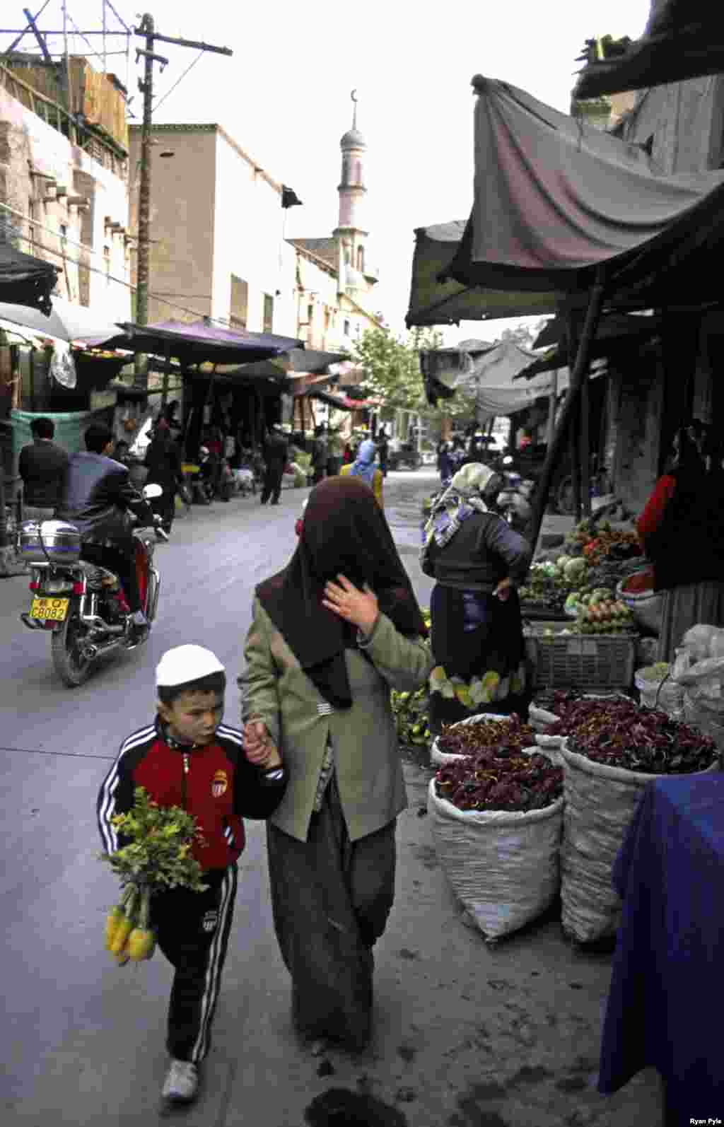 A woman, fully veiled, walks down a street with her son. - Street vendors in Kashgar sell hand-made candy, fresh mutton, or hand-sewn skull caps. Donkey-cart drivers navigate the narrow streets.